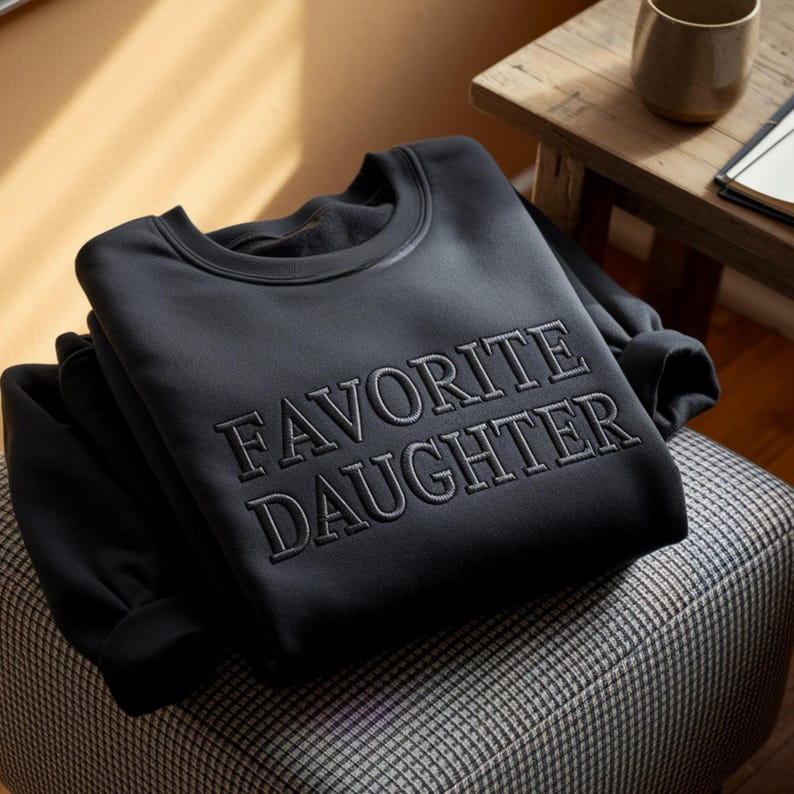Puede incluir: Sudadera negra con el texto bordado "FAVORITE DAUGHTER". La sudadera de cuello redondo est&aacute; doblada sobre un otom&aacute;n estampado. Una peque&ntilde;a mesa de madera y una taza est&aacute;n en el fondo.