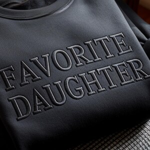 Puede incluir: Una sudadera negra con el texto bordado "FAVORITE DAUGHTER" en un tono gris m&aacute;s claro. La sudadera est&aacute; doblada y la tela tiene una textura suave. El dise&ntilde;o es simple y elegante, adecuado para ropa casual.