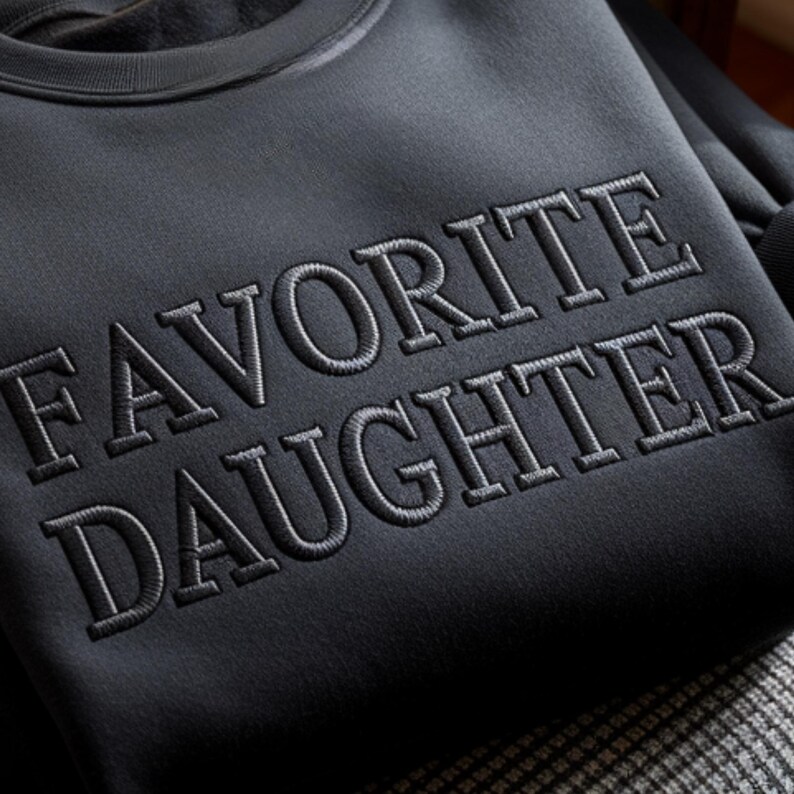 Puede incluir: Una sudadera negra con el texto bordado "FAVORITE DAUGHTER" en un tono gris m&aacute;s claro. La sudadera est&aacute; doblada y la tela tiene una textura suave. El dise&ntilde;o es simple y elegante, adecuado para ropa casual.