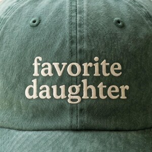 Pode incluir: Bon&eacute; de beisebol verde com o texto bordado "favorite daughter" em branco. O bon&eacute; tem uma aba curva e um design cl&aacute;ssico de seis pain&eacute;is. O tecido tem uma apar&ecirc;ncia ligeiramente desbotada.