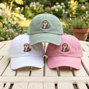 Puede incluir: Tres gorras de béisbol en verde salvia, blanco y rosa empolvado. Cada gorra presenta un bordado de una nutria marrón sentada en agua azul en el panel frontal. Las gorras tienen una corona estructurada, una visera curva y un botón superior.