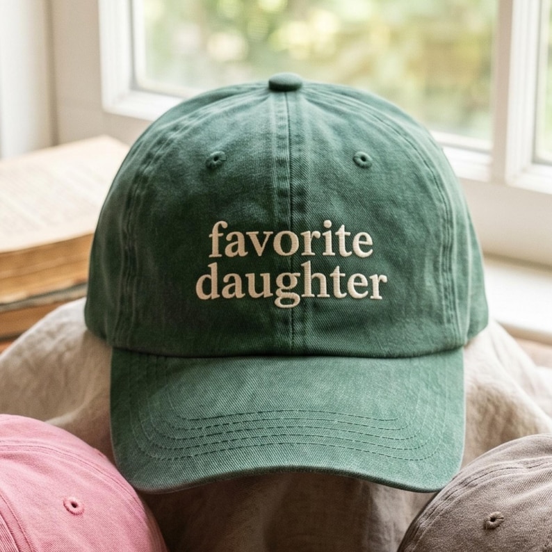 May include: A green baseball cap with the embroidered text "favorite daughter" in white. The cap has a curved brim and a fabric strap closure. The hat is made of a soft, washed cotton material.