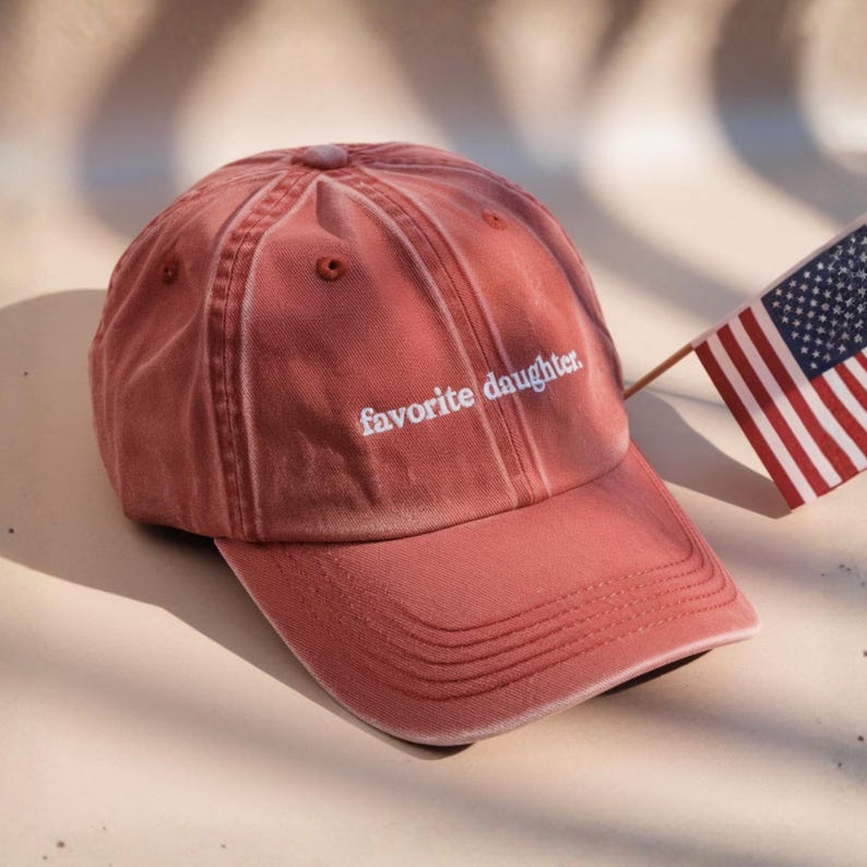 K&ouml;nnte beinhalten: Staubrosa Baseballkappe mit dem wei&szlig;en Schriftzug "favorite daughter". Die Kappe hat einen gebogenen Schirm und ein klassisches sechsteiliges Design. Im Hintergrund ist eine amerikanische Flagge zu sehen.