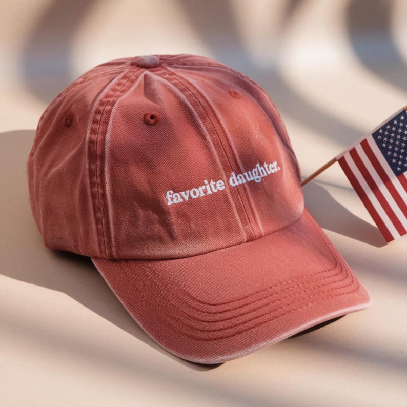 K&ouml;nnte beinhalten: Verblasste rosafarbene Baseballkappe mit dem wei&szlig;en Schriftzug "favorite daughter". Die Kappe hat einen gebogenen Schirm und einen Stoffriemenverschluss. Im Hintergrund ist eine amerikanische Flagge zu sehen.