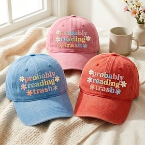 May include: Three baseball caps in pink, blue, and red, each featuring the embroidered text "probably reading trash" in multi-colored letters, accented with small embroidered daisy flowers in various pastel colors on the front panels.