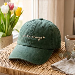 May include: A dark green baseball cap featuring the embroidered text "you are enough" in a light cream cursive font, accompanied by a small heart symbol. The cap has a structured crown, a curved brim, and visible stitching details on the fabric.
