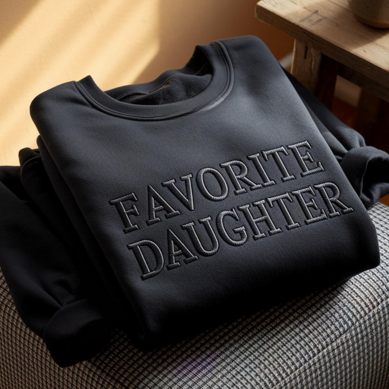 Puede incluir: Una sudadera negra doblada con el texto bordado "FAVORITE DAUGHTER" en un hilo gris oscuro a juego. La sudadera est&aacute; hecha de un material suave y est&aacute; doblada cuidadosamente sobre una superficie estampada.