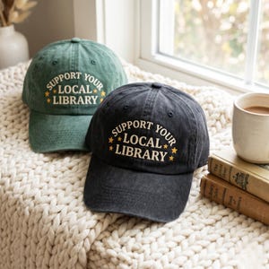 May include: Two baseball caps, one dark gray and one green, with the text "SUPPORT YOUR LOCAL LIBRARY" embroidered in white and yellow. The caps are sitting on a cream-colored, textured blanket.