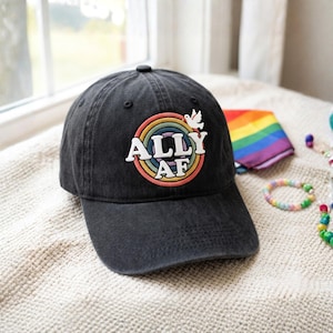 May include: A charcoal gray baseball cap featuring an embroidered circular patch with concentric rainbow rings. The text ALLY AF is stitched in white letters across the center, accompanied by a small white dove icon positioned at the top right.