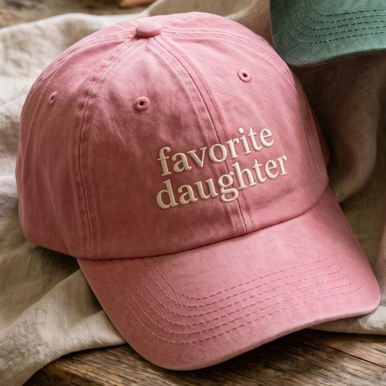May include: A pink baseball cap with the embroidered text "favorite daughter" in white. The cap has a curved brim and a six-panel design. The hat is made of a soft, textured fabric.