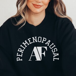May include: Black crewneck sweatshirt with a white, arch-shaped graphic that says 'PERIMENOPAUSAL AF'. The sweatshirt is shown on a model against a white background.