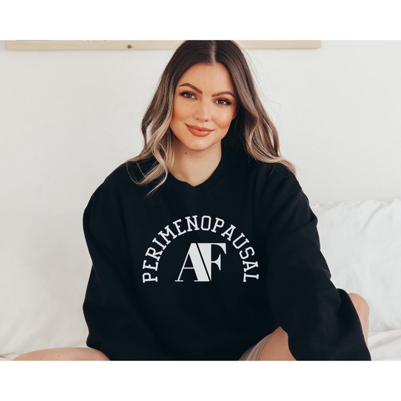 May include: Black crewneck sweatshirt with a white, arch-shaped graphic that says 'PERIMENOPAUSAL AF'. The sweatshirt is shown on a model against a white background.
