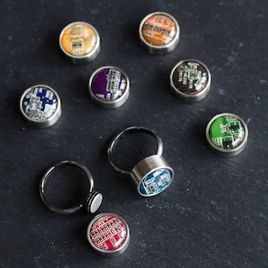 Peut inclure: Une collection de bagues et de bijoux circulaires avec des motifs de circuits imprimés colorés. Les bagues ont des anneaux argentés et les pièces circulaires présentent diverses couleurs, notamment orange, violet et vert. Les bijoux sont posés sur un fond sombre.