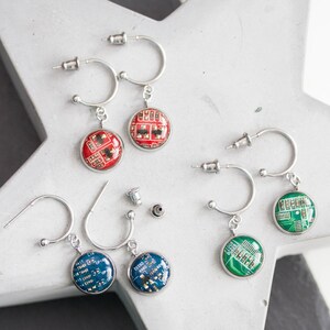 May include: Three pairs of silver hoop earrings with a round pendant. Each pendant features a different coloured circuit board design. The circuit boards are red, green, and blue.