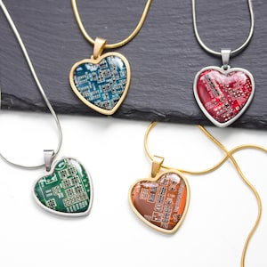 May include: Heart-shaped pendant necklaces with circuit board designs. The pendants are in various colors, including blue, red, green, and brown, with gold and silver-toned chains. The necklaces are displayed on a dark gray surface.
