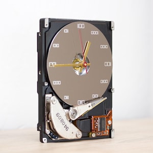 May include: A wall clock constructed from a repurposed hard drive. The circular silver platter features binary code numerals in place of standard clock numbers. It includes gold-colored hour and minute hands and a thin red second hand.