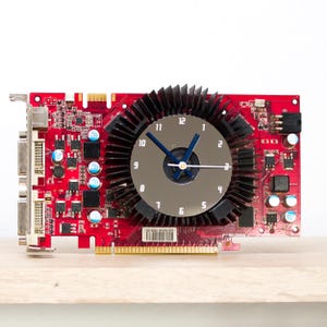 May include: A red computer graphics card repurposed as a clock. The clock face is gray with black numbers and blue hands. The clock is mounted on the graphics card's circuit board, which has various electronic components.