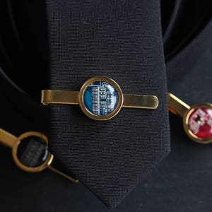 Pode incluir: Uma presilha de gravata dourada com um design circular de placa de circuito azul e branco. A presilha de gravata está em uma gravata preta, com duas outras presilhas de gravata visíveis no fundo.