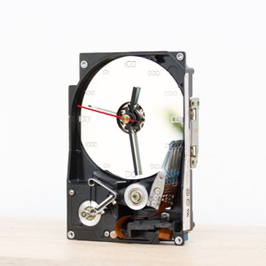 Puede incluir: Un reloj único hecho de un disco duro de computadora reutilizado. La esfera del reloj es blanca con números negros y manecillas negras, con un segundero rojo. La carcasa del disco duro es negra y el reloj está sobre una superficie clara.