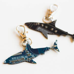 May include: Two shark-shaped keychains crafted from recycled circuit boards. One keychain features a dark blue printed circuit board with visible electronic components, while the other is black. Both include silver-toned metal lobster claw clasps.
