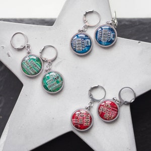 Peut inclure: Trois paires de boucles d'oreilles pendantes circulaires composées de segments de circuits imprimés verts, bleus et rouges dans des montures en métal argenté avec fermoirs à levier. Chaque boucle montre des composants électroniques et du texte.