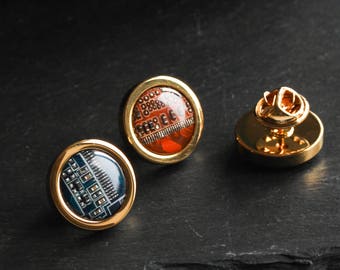 May include: Three gold-coloured lapel pins featuring circuit board designs. Two pins have a round, domed design with a gold border, one with a blue and green circuit board and the other with a red and orange circuit board. The third pin has a gold post.