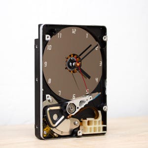 Può includere: Un orologio unico realizzato con un disco rigido di computer riciclato. Il quadrante è di colore marrone chiaro con lancette nere e numeri bianchi. L'involucro dell'orologio è nero e i componenti interni del disco rigido sono visibili.