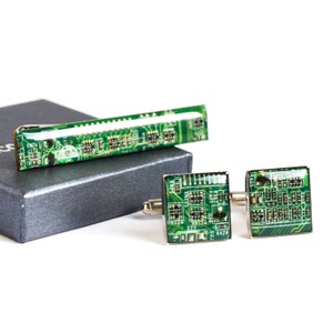 Puede incluir: Un juego de gemelos cuadrados y un pasador de corbata rectangular con diseño de placa de circuito verde. El conjunto se presenta en una caja gris oscuro, ideal para los amantes de la tecnología y la electrónica.