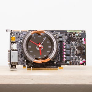 May include: A black computer graphics card with a copper cooling fan that has been modified to include a clock face. The clock face has red hands and a white second hand.