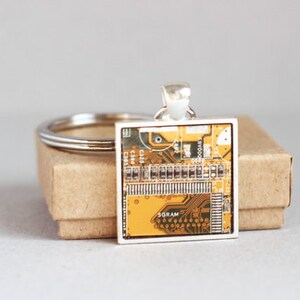 May include: Square pendant featuring a circuit board design in shades of yellow, brown, and green, set in a silver-tone frame. The pendant is attached to a silver keyring, resting on a small, brown cardboard box.