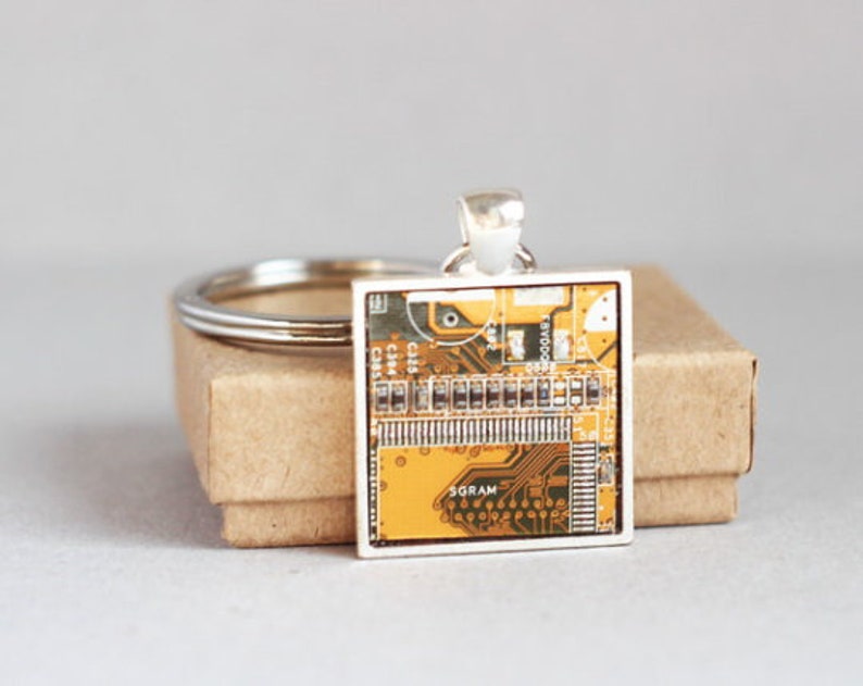 May include: Square pendant featuring a circuit board design in shades of yellow, brown, and green, set in a silver-tone frame. The pendant is attached to a silver keyring, resting on a small, brown cardboard box.