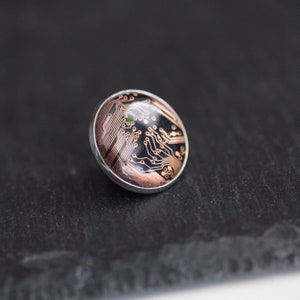 Op de afbeelding: Een ronde, zilverkleurige pin met een zwart en koperkleurig printplaatontwerp.