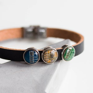 May include: A black leather bracelet with three circular charms featuring colorful circuit board designs. The bracelet has a silver clasp and a light brown leather backing. The charms are blue, yellow, and green.