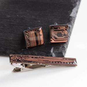 Könnte beinhalten: Ein Set aus drei silbernen Manschettenknöpfen und einem Krawattenclip mit einem kupferfarbenen Leiterplatten-Design.