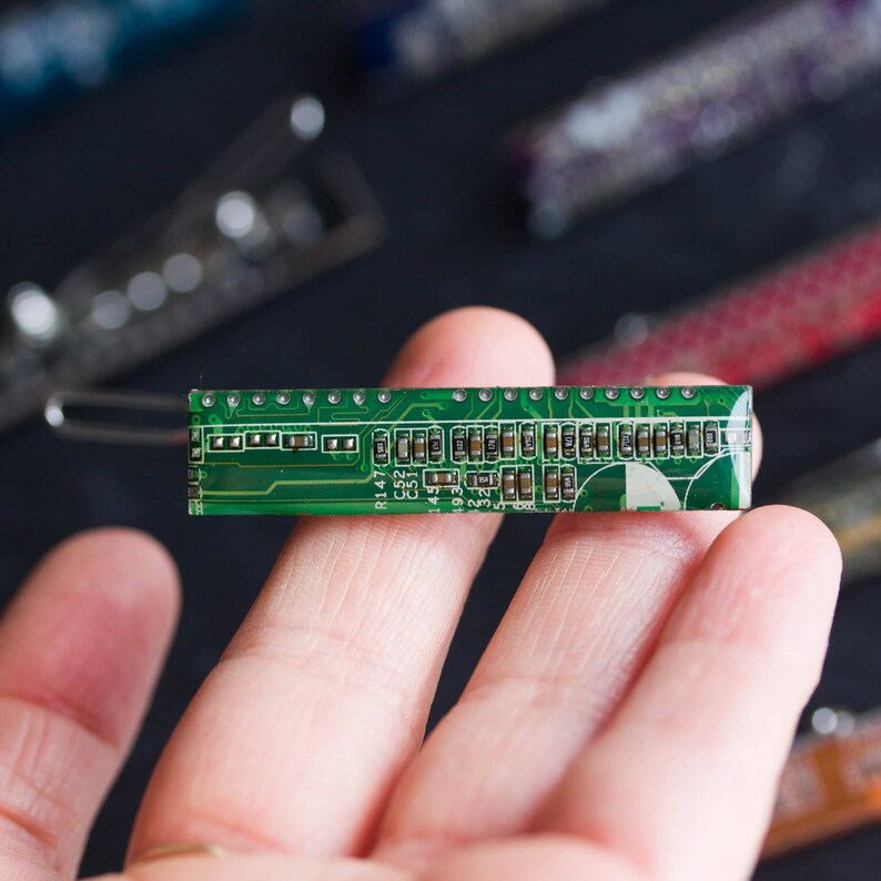 May include: A green circuit board hair clip with a clear, curved barrette clasp. The clip features a detailed electronic component design with various small chips and connectors. Other hair clips are visible in the blurred background.