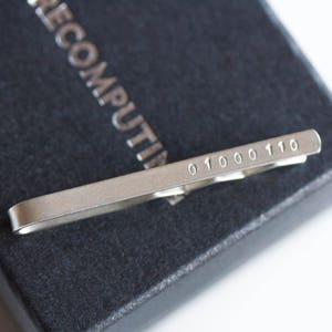 Pode incluir: Uma barra de gravata prateada com o código binário "01000110" gravado. A barra de gravata está sobre uma superfície texturizada cinza escuro. A palavra "RECOMPUTING" está em relevo na superfície.