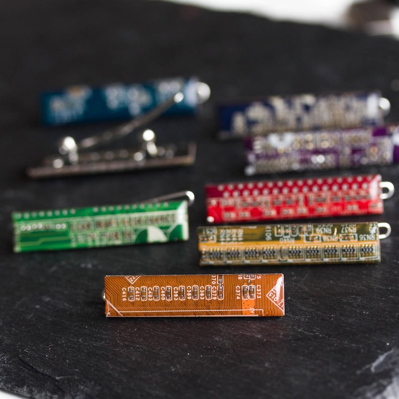 May include: A collection of colorful circuit board tie clips and a tie bar. The tie clips feature various colors, including green, red, and blue, with intricate electronic component patterns. The tie bar is silver. The items are displayed on a dark surface.