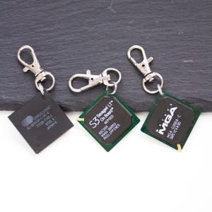 Op de afbeelding: Drie vierkante computerchip-sleutelhangers met zilverkleurige metalen karabijnsluitingen. De chips bevatten gedrukte tekst zoals Cirrus Logic CL-605436-1-QC-C, S3 Savage4 LT On Board en Matrox MGA-G200A-C op een donkergroene of zwarte ondergrond.