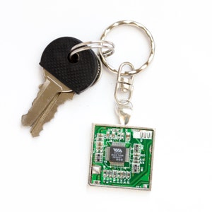 May include: A silver key ring with two keys and a square pendant. The pendant features a green circuit board design with a silver frame. The keys have a black rubber grip.