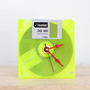 Puede incluir: Un reloj de disquete amarillo con una etiqueta plateada que dice "imation 2HD IBM formateado: 1,44 MB". El reloj tiene manecillas rojas y una manecilla de segundos negra.