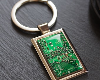 May include: A rectangular silver keychain with a green circuit board design encased in clear resin. The keychain has a silver ring for attaching keys. The circuit board features intricate electronic components.