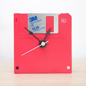 May include: A red floppy disk clock with black clock hands and a silver label. The label reads "3M FORMATTED/FORMATEES IBM DS,HD 1.44 Mb/1.44 Mo". The clock hands are black and the clock face is a bright red color.
