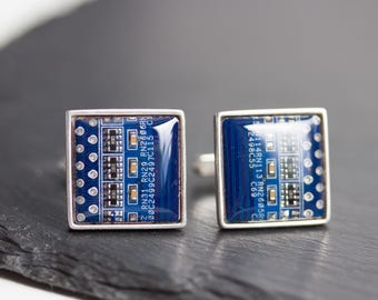 May include: A pair of square cufflinks with a silver frame. Each cufflink has a blue circuit board design with small electronic components and white text. The cufflinks are presented on a dark grey surface.