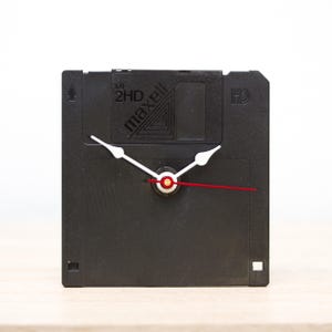 Puede incluir: Un reloj de disquete negro con agujas blancas y una aguja de segundos roja. El disco está etiquetado como "Maxell 2HD MF".