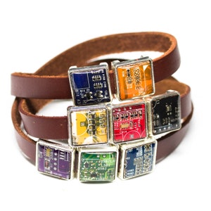 Puede incluir: Una pulsera envolvente de cuero marrón con múltiples colgantes cuadrados. Cada colgante es una placa de circuito en miniatura encapsulada en un marco plateado. Las placas de circuito muestran varios colores y componentes electrónicos, creando un accesorio único e inspirado en la tecnología.
