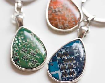 May include: Three teardrop-shaped keychains with silver metal frames. Each keychain features a different colored epoxy resin filled with a circuit board pattern. The colors are green, blue, and orange.