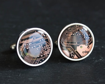 May include: A pair of silver-toned cufflinks with a circuit board design. The round cufflinks feature a black background with copper-colored circuit patterns, encased in a silver border. The cufflinks are displayed on a dark surface.