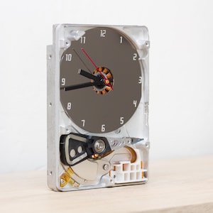 May include: A wall clock constructed from a repurposed hard drive, featuring a circular metallic platter as the clock face with printed numbers one through twelve. The clock includes black hour and minute hands and a thin red second hand.