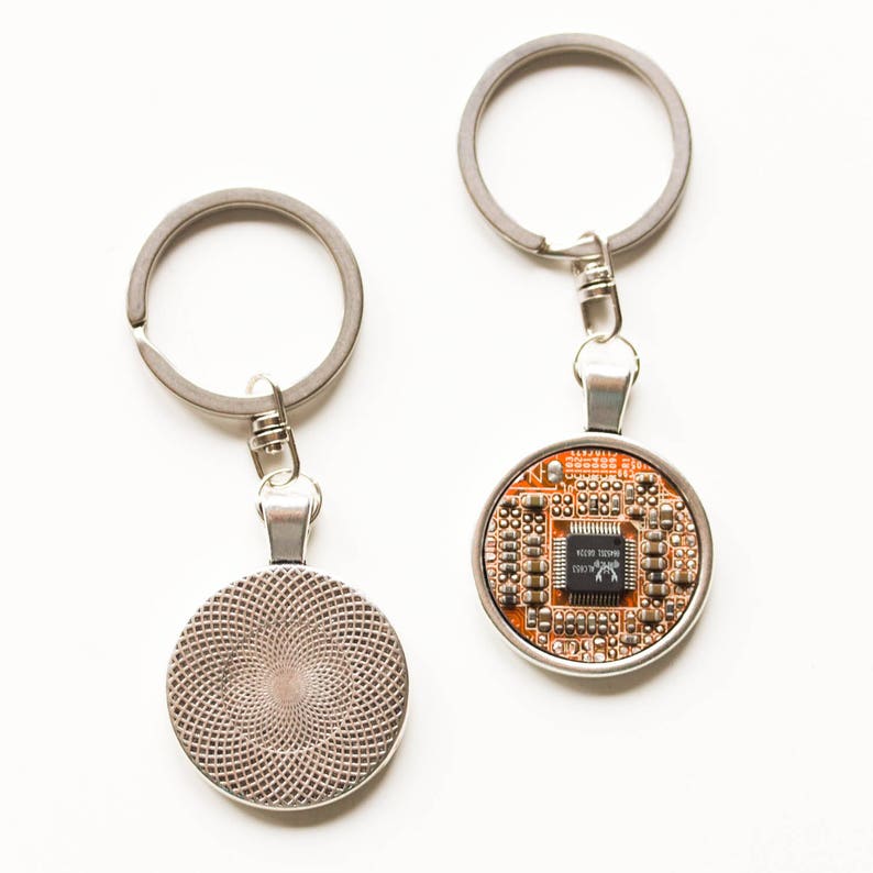 May include: Two keychains with silver-colored rings and pendants. One pendant displays a circuit board design with orange, brown, and black components. The other pendant has a textured, silver-colored back. The keychains are on a white background.