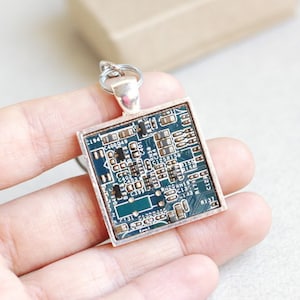 May include: Square pendant featuring a section of a circuit board encased in a silver-toned frame. The circuit board has a teal background with various electronic components. The pendant is attached to a silver-toned bail.